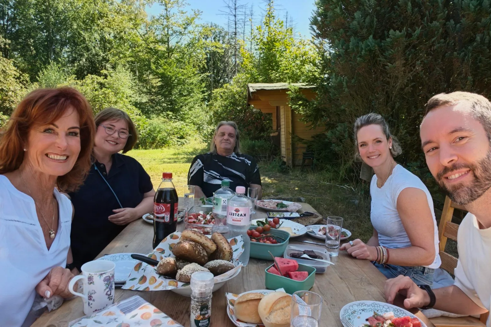 Team an einer langen Holztafel beim Sommerfest, Wald und Wiese im Hintergrund
